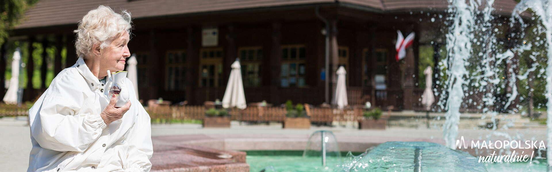 Eine ältere Frau sitzt an einem Brunnen in Wysowa Zdrój und hält einen Trinkbecher für Mineralwasser in der Hand.